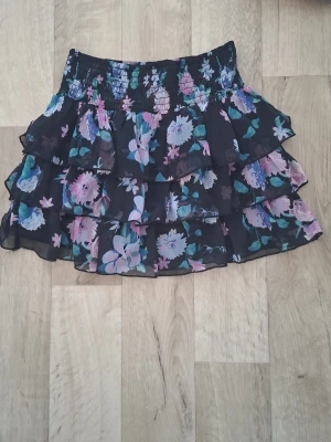 Blommig volangkjol från Lindex - Söt kortkjol från Lindex med blommigt mönster i rosa, lila, blått och grönt på svart bakgrund. Kjolen har flera volanger och smockad midja som ger en bekväm passform. Perfekt för dig som gillar färg och mönster!