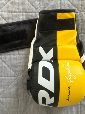 RDX MMA handskar svart/gul/vit - Säljer ett par RDX MMA handskar i svart, gul och vit färg med bred kardborreknäppning och öppna fingrar. Tillverkade i slitstarkt syntetläder och har tryckt logotyp samt signatur på ovansidan. Perfekta för träning och sparring. Använt en gång!