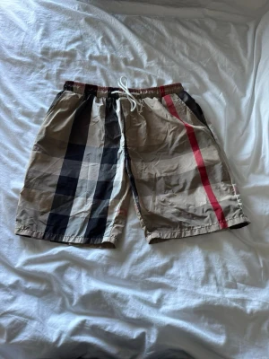 Beige rutiga shorts/badshorts - Säljer ett par beige shorts/badshorts med klassiskt rutigt mönster i svart, rött och vitt. Shortsen har elastisk midja med vit snörning och är tillverkade i ett lätt och bekvämt material. Perfekta för sommaren och har en avslappnad passform.