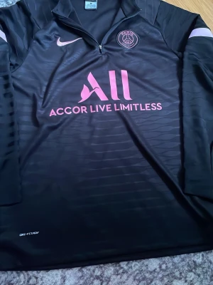 Svart PSG träningsjacka Nike M - Säljer en svart långärmad träningsjacka från Nike med PSG-logga och rosa detaljer. Jackan har halv dragkedja, Dri-Fit material och tryck med 'Accor Live Limitless' på bröstet. Perfekt för fotbollsträning eller sport.