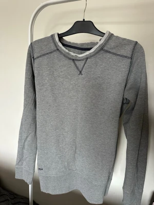 Grå sweatshirt från ARTB - En stilren grå sweatshirt från ARTB med mörkblå sömmar och broderad logga i nacken. Tröjan har rund halsringning med rå kant och långa ärmar. Perfekt för en avslappnad och trendig look.