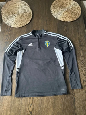 Svart Adidas Sverige träningströja - Svart långärmad träningströja från Adidas med Sveriges landslagsemblem på bröstet. Tröjan har vita detaljer längs sidorna och axlarna samt dragkedja vid halsen. Perfekt för fotbollsträning eller sportiga dagar.