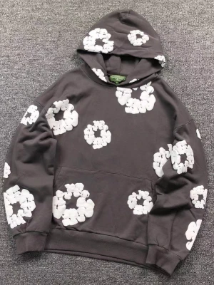Denim tears  hoodie med vita blommor - Cool mörkgrå hoodie med stora vita blommor tryckta över hela plagget. Hoodien har en rymlig huva och klassisk känguruficka framtill. Perfekt för dig som gillar unika mönster och avslappnad stil.