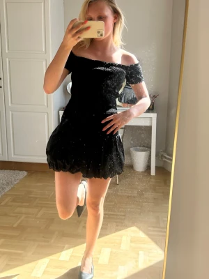 Svart offshoulder kort klänning💖💖 - Trendig svart kort klänning med offshoulder-design och puffig kjol. Klänningen har en smockad överdel som ger en snygg passform och är tillverkad i ett glittrigt material som fångar ljuset. Perfekt för dig som vill sticka ut och känna dig extra stylish.