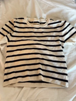 Randig kortärmad topp i bomull - Säljer en vit och svart randig topp med korta ärmar och rund hals. Toppens material är mjuk bomull och den har en klassisk, normal passform. Perfekt för dig som gillar enkel och stilren design.