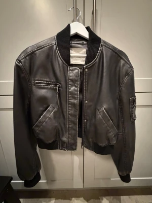 Svart bomberjacka i skinn - Snygg svart bomberjacka i skinn med ribbade muddar och krage. Jackan har dragkedja framtill, två snedställda fickor med lock, en bröstficka med dragkedja och en ärmficka med dragkedja. Klassisk och stilren modell som passar till det mesta.