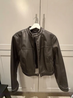 Svart cropped skinnjacka Gina Tricot - Snygg svart cropped skinnjacka från Gina Tricot med hög krage och två knappar. Jackan har markerade sömmar, dragkedja framtill och två fickor. Perfekt för dig som vill ha en trendig och stilren look.