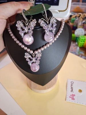 Ljuslila pärlhalsband med fjäril - Säljer ett halsband med ljuslila pärlor och matchande hänge i form av en glittrig fjäril med en stor lila pärla. Halsbandet har en elegant och söt design, perfekt för dig som gillar detaljer och pastellfärger. Matchande örhängen medföljer.