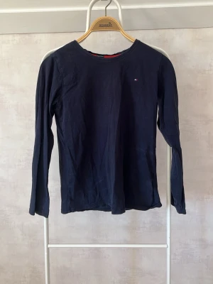 Mörkblå långärmad tröja Tommy Hilfiger - En stilren mörkblå långärmad tröja från Tommy Hilfiger med rund halsringning och liten logga på bröstet. Tillverkad i mjuk bomull som känns skön mot huden. Perfekt till jeans eller kjol för en clean look.