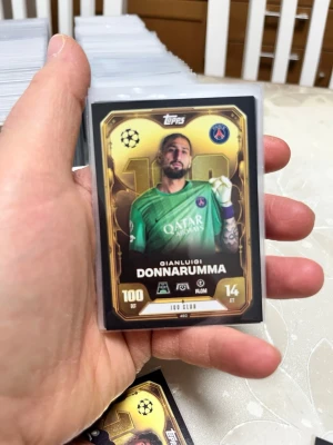 Topps Match Attax Donnarumma-kort PSG - Samlarkort från Topps Match Attax med Gianluigi Donnarumma i PSG, 100 Club-version. Kortet har guldfärgad ram, PSG-logga och detaljer i svart och vitt. Perfekt för dig som gillar fotboll och samlarkort.
