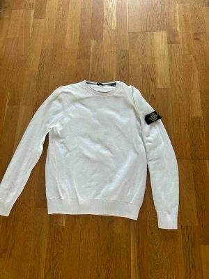 Vit stickad tröja från Stone Island - Vit stickad tröja från Stone Island med klassisk rund hals och ribbade muddar. Tröjan har den ikoniska Stone Island-loggan på vänster ärm. Perfekt för dig som gillar stilrena och trendiga plagg.