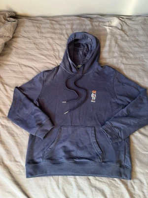 Blå hoodie från Polo Ralph Lauren - Säljer en klassisk marinblå hoodie från Polo Ralph Lauren med den ikoniska björnen broderad på bröstet. En tidlös hoodie i skönt material. Viktig information om storlek: • Tag: Storlek L (Large). • Passform: Tröjan har krympt i tvätt/tork och sitter nu som en XS. Se därför till att utgå från att den är liten i storleken! Skick: Tröjan är i bra skick överlag och har kvar sin fina färg, men det finns en skada vid huvan/halsen (se bild 3). Det är två mindre hål där dragsnörena suttit, men detta är 