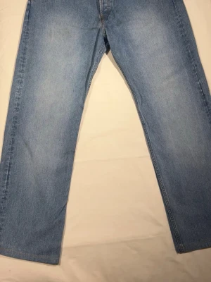 Blå raka jeans i klassisk modell - Säljer ett par ljusblå jeans med rak passform och klassisk femficksdesign. Jeansen har en enkel look utan slitningar och är tillverkade i denim. Perfekta för dig som gillar en tidlös och avslappnad stil.