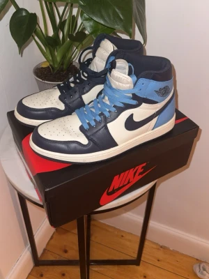 Nike Air Jordan 1 High blå/vit - Säljer ett par Nike Air Jordan 1 High sneakers med klassisk design. Skorna har en kombination av blått, ljusblått och vitt läder, ikonisk Swoosh på sidan och högt skaft. Perfekta för dig som vill ha en stilren och sportig look.