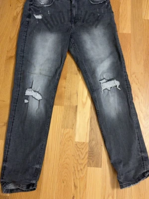 Grå slitna jeans från Hoodrich - Säljer ett par grå jeans från Hoodrich med coola slitningar och tryck på framsidan. Jeansen har fem fickor, texten 'FROM NOTHING TO SOMETHING' på insidan av linningen och är tillverkade i denim. Perfekt för dig som gillar streetstyle och en avslappnad look.