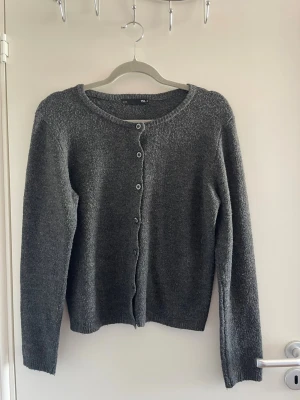 Mörkgrå kofta från H&M - En enkel och stilren mörkgrå kofta från H&M med rund halsringning och knappar framtill. Koftan är stickad och har långa ärmar samt ribbade muddar vid ärmslut och nederkant. Perfekt att slänga på sig när det är kyligt.