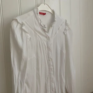 Vit sommarblus med volang från Tissaia - Säljer en söt vit blus från Tissaia med volangdetaljer vid krage och axlar. Blusen har knappar framtill och lång ärm. Perfekt för sommaren och fina middagar. Storlek s