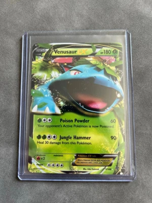 Venusaur EX Pokémonkort - Säljer ett Venusaur EX samlarkort från Pokémon, med holografisk yta och färgstark design. Kortet har attacker som Poison Powder och Jungle Hammer och är inplastat i ett skyddsfodral. Perfekt för dig som samlar eller spelar Pokémonkort.