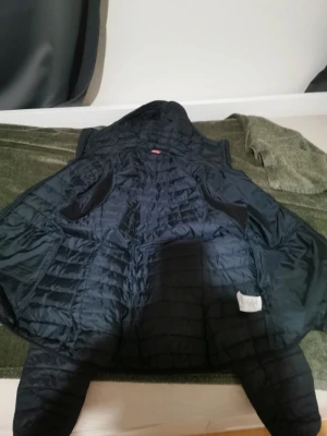 Svart dunjacka med huva från Haglöfs - Säljer en svart dunjacka från Haglöfs med quiltad design och huva. Jackan har diskret logga på ärmen och är tillverkad i lätt, isolerande material som håller dig varm under kalla vinterdagar. Perfekt för dig som vill ha en stilren och funktionell jacka.