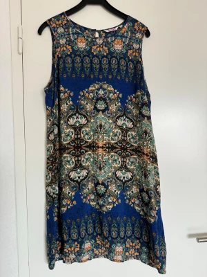 Blå ärmlös klänning med mönster - Säljer en blå ärmlös klänning med ett detaljerat orientaliskt mönster i grönt, vitt och brunt. Klänningen har rund halsringning och rak passform, perfekt för dig som gillar färgstarka och unika plagg.