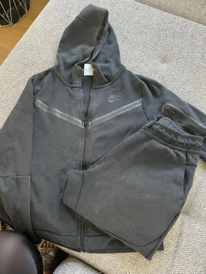 Svart Nike Tech - Svart set från Nike i färg med matchande hoodie. Väl använt och små skavanker. Strl L  147/158