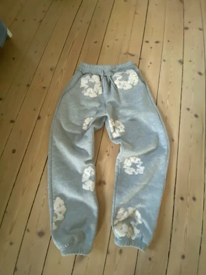 Grå mjukisbyxor från denim tears  - Säljer ett par grå mjukisbyxor från denim tears med vita blomliknande tryck på benen. Byxorna har resår i midjan och vid benslut, vilket ger en avslappnad och skön passform. Perfekta för chill dagar och häng med kompisar.