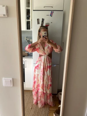 Blommig långklänning med cut-outs - Säljer en långklänning med blommigt mönster i rosa och vitt. Klänningen har v-ringning, vida ärmar och snygga cut-outs i midjan. Använd max 2 gånger, helt ny skick.
