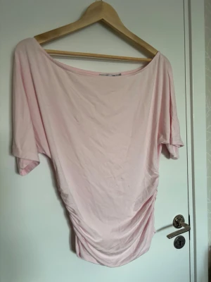 Ljusrosa topp från lager 157 - Ljusrosa topp med rynkade detaljer längs sidorna. Modellen har en bred, rund halsringning och korta ärmar. Materialet är mjukt och stretchigt. Från lager 157