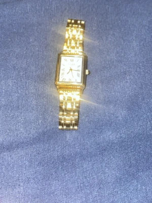 Guldfärgad klocka från Regal - Stilren guldfärgad klocka från Regal med fyrkantigt ur och vit urtavla med romerska siffror. Länken är metall och har en klassisk design som känns både elegant och tidlös. Perfekt för dig som vill ha en snygg accessoar som sticker ut. Säljer den för den kommer inte till användning. Hör av dig gärna så disskuterar vi priset 