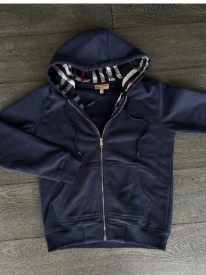 Marinblå hoodie från Burberry - Snygg marinblå hoodie från Burberry med dragkedja och klassiskt rutigt foder i huvan och längs insidan. Hoodien har ribbade muddar, fickor framtill och justerbar huva med snören. Perfekt för dig som gillar stilrena och exklusiva plagg.