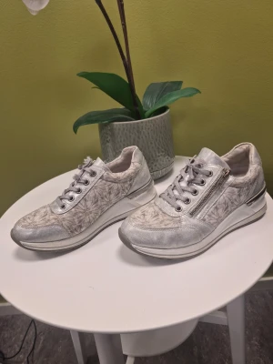 Rieker Remonte Sneakers med kilklack D3203-90 silver Storlek 36 - Snygga sneakers från Rieker i ljusgrått med subtilt blommönster och silverdetaljer. Skorna har snörning och en praktisk dragkedja på sidan. Ovandelen är i textil och syntet, sulan är vit med grå inslag. Perfekt för dig som gillar stilrena och unika sneakers.