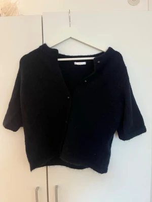 Mörkblå kofta med kort ärm - Mysig svart kofta med korta ärmar och rund hals. Koftan har knäppning framtill och är från gina tricot. Oanvänd. 