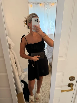 Svart kort klänning med knytband - Säljer en svart kort klänning med smala axelband som knyts på axlarna. Klänningen har en enkel och stilren design med lätt volang upptill och är perfekt för dig som gillar en feminin look. Materialet känns lätt och bekvämt.