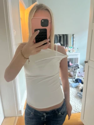 Vit oneshoulder topp - Snygg vit oneshoulder topp med tight passform. Toppen är ärmlös och har en stilren, minimalistisk look som passar perfekt till jeans eller kjol. Materialet känns mjukt och stretchigt för en bekväm känsla.