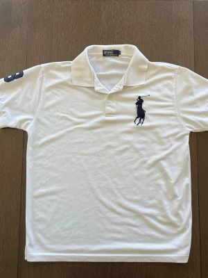 Vit pikétröja från Polo Ralph Lauren - Vit pikétröja från Polo Ralph Lauren med klassisk krage och korta ärmar. Tröjan har en broderad logotyp med en polospelare på bröstet och siffran 3 på ena ärmen. Perfekt för en sportig och stilren look.