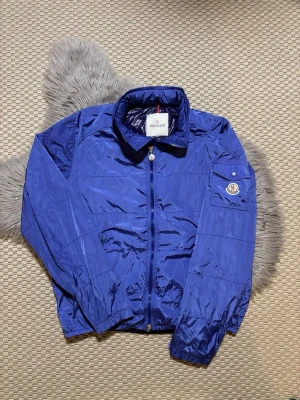 Blå vindjacka från Moncler (M/L) - Snygg blå vindjacka från Moncler med klassisk krage och dragkedja framtill. Jackan har en ficka med Moncler-logga på ena ärmen och elastiska muddar vid ärmslut och nederkant. Materialet är glansigt och lätt, perfekt för att hålla vinden ute.