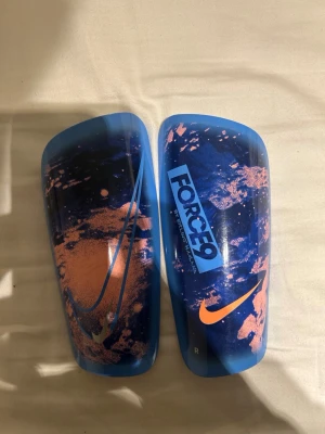 Nike benskydd blå/orange FORCE9 - Säljer ett par Nike benskydd i blått med orange detaljer och FORCE9-tryck. Snyggt mönster med stänk-effekt och tydliga Nike-loggor. Perfekta för fotbollsträning eller match. Lätta och smidiga att använda.