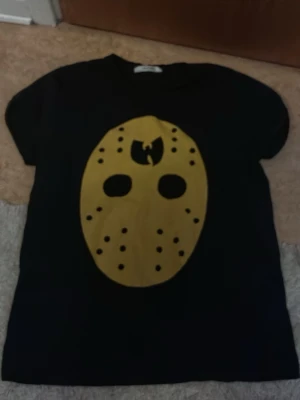Wu-Tang Clan-loggan på tröjan  - Cool svart t-shirt från Best med ett stort gult tryck av en mask på framsidan, inspirerad av Wu-Tang Clan-loggan. T-shirten har korta ärmar och en klassisk passform. Perfekt för dig som gillar streetwear och unika prints.
