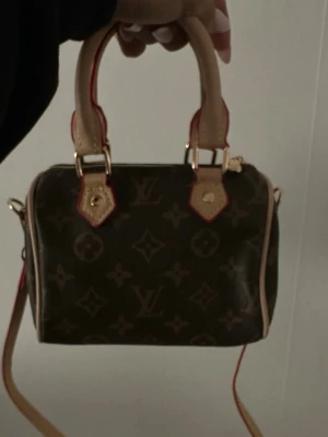 Monogram handväska - Säljer en klassisk handväska i brunt monogrammönster med beigea handtag och detaljer i rött. Väskan har guldfärgade metalldetaljer och avtagbar axelrem. Perfekt storlek för det viktigaste och ikonisk design som alltid känns rätt.