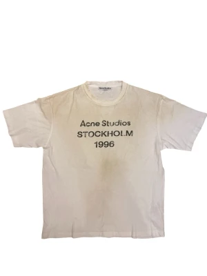 Vit Acne Studios t-shirt Stockholm - Vit t-shirt från Acne Studios med svart text 'Acne Studios STOCKHOLM 1996' på bröstet. Klassisk rund halsringning och korta ärmar. Tillverkad i mjuk bomull för en skön och avslappnad look. Dessa är oversized så den sitter som en herr medium.