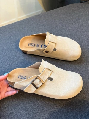 Beige Birkenstock tofflor - Säljer ett par klassiska beige tofflor från Birkenstock i mjuk mocka. Modellen har en bred rem med metallspänne och öppen häl. Sulan är ergonomiskt utformad för extra komfort. Perfekta för dig som gillar stilrena och bekväma skor. Priset kan diskuteras 