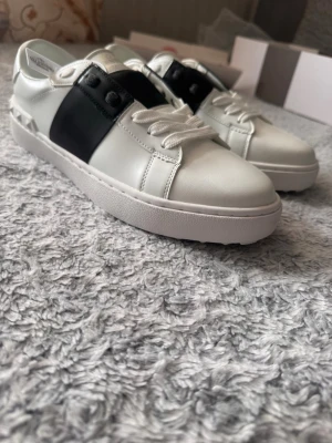 Valentino skor, storlek 43 - Snygga vita sneakers från Valentino med en bred svart läderdetalj över sidan. Skorna har klassisk låg profil, vita snören och en robust vit sula. Perfekt för dig som gillar stilrena och trendiga skor med exklusiv känsla.