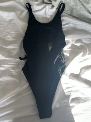 Svart baddräkt med snörning - Säljer en svart baddräkt med smala axelband och snörningsdetaljer på sidorna. Baddräkten har en klassisk rund halsringning och justerbara axelband. Materialet ser ut att vara ribbat och stretchigt, perfekt för strand eller pool.