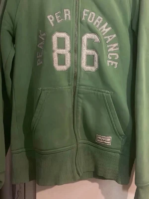 Grön hoodie från Peak Performance - Snygg grön hoodie från Peak Performance med dragkedja framtill och stor vit 86-print på bröstet. Hoodien har två fickor fram och ribbade muddar vid ärmslut och nederkant. Perfekt för en avslappnad och sportig stil.