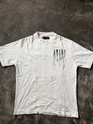 Vit AMIRI t-shirt med broderad logga - Säljer en vit t-shirt från AMIRI med broderad logga och färgstänk-effekt på bröstet. T-shirten har korta ärmar, rund halsringning och är gjord i mjuk bomull. Tröjan har aldrig varit använd och pris kan diskuteras via snabb affär.