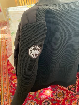 Svart dunväst från Canada Goose - Snygg svart dunväst från Canada Goose med quiltad framsida och stickade ärmar. Västen har hög krage, två dragkedjefickor och ett tydligt Canada Goose-märke på ärmen. Perfekt för kalla dagar och stilrena outfits.