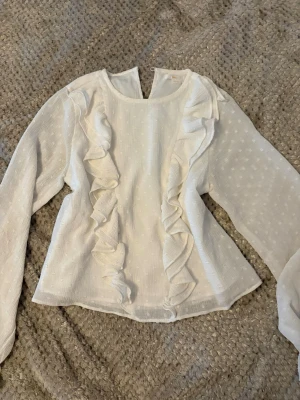 Vit blus med volanger och prickar - Säljer en vit blus med långa ärmar och dekorativa volanger framtill. Blusen har ett subtilt prickigt mönster och är tillverkad i ett lätt, transparent material som ger en luftig känsla. Rund halsringning och knapp i nacken för enkel på- och avtagning.
