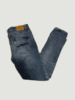 Blå jeans från Nudie Jeans - Säljer ett par klassiska blå jeans från Nudie Jeans med snyggt slitna detaljer och broderad bakficka. Jeansen har rak passform och fem fickor, tillverkade i bomull. Perfekta för dig som gillar en tidlös och cool look.