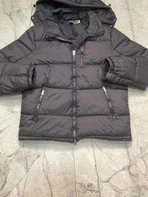 Svart pufferjacka från Polo Ralph Lauren - Säljer en svart pufferjacka från Polo Ralph Lauren med huva och två dragkedjeförsedda fickor framtill. Jackan har en röd broderad logga på bröstet och är vadderad för extra värme. Perfekt för kalla vinterdagar och riktigt snygg streetstil.