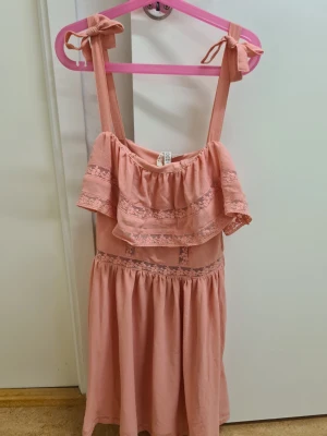 Rosa klänning med volang från H&M 32 - Supersöt rosa klänning från H&M Divided med knytband på axlarna och volang upptill. Klänningen har spetsdetaljer och är gjord i mjuk bomull, perfekt för varma dagar. Den är kort och har en luftig passform som känns både bekväm och stylish.